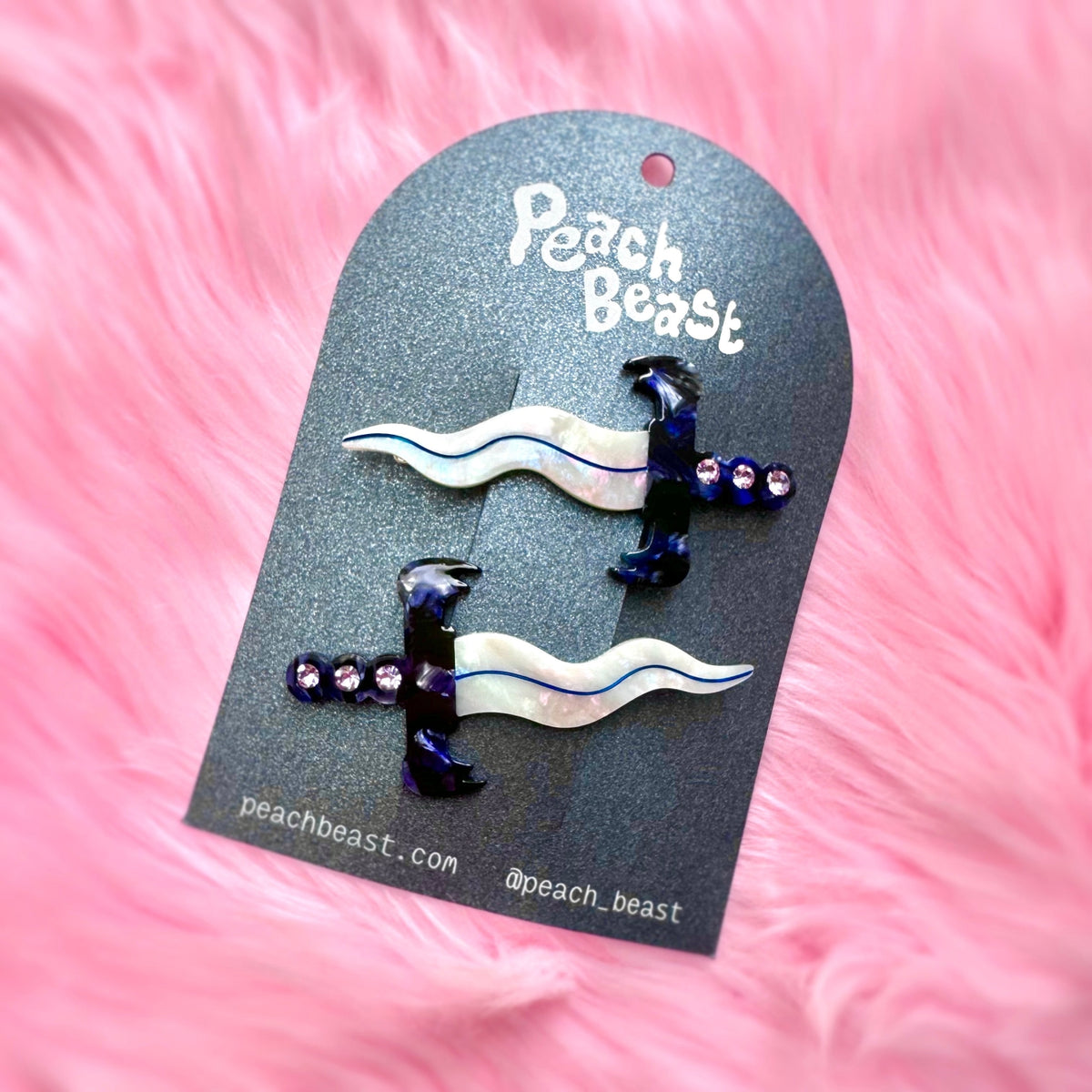 Two swords with sqiggly iridescent blades featuring blue hilts and purple rhinestones on a black backing card against a pink background.
