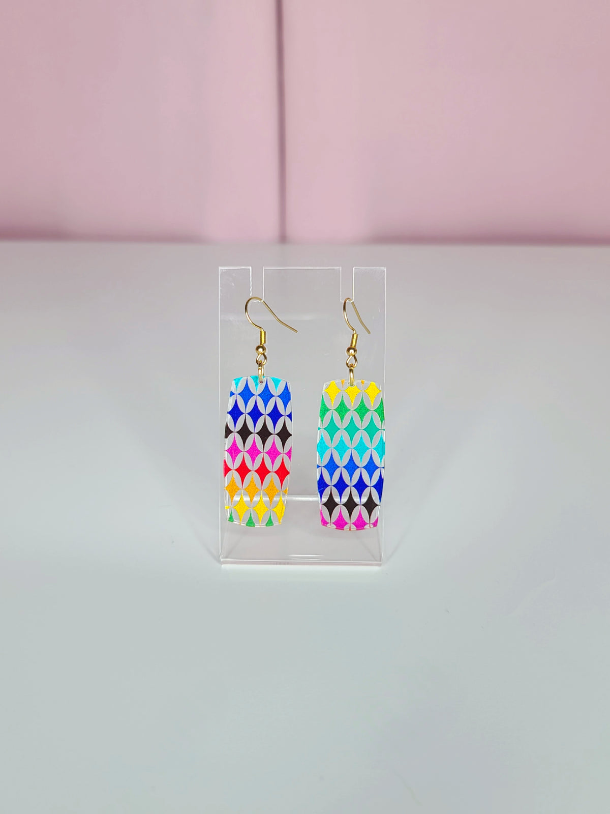 Rainbow Harlequin Dream Earrings with a colorful geometric pattern, displayed on a clear stand against a pink and white background.