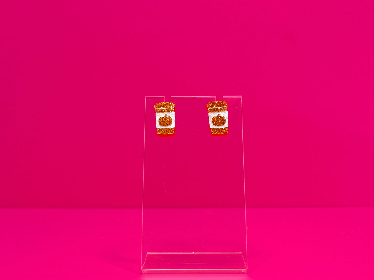 Orange flitter pumpkin spice cup earrings on a clear stand against a pink background.
