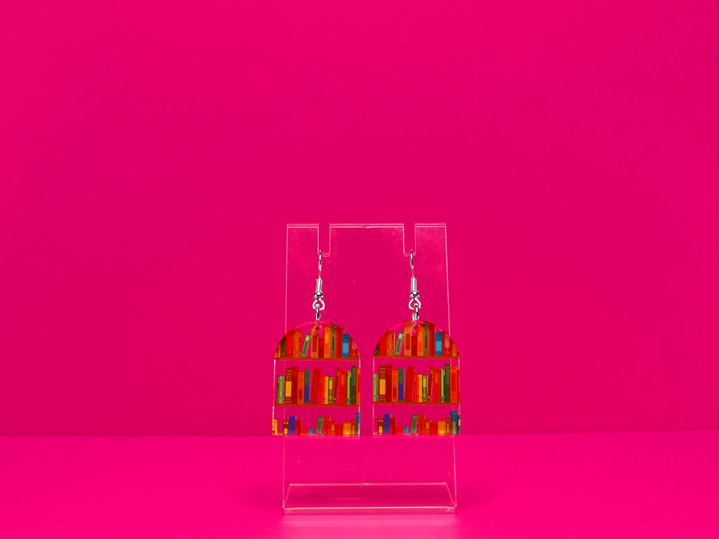 Arched bookshelf earrings on a clear stand against a pink background.