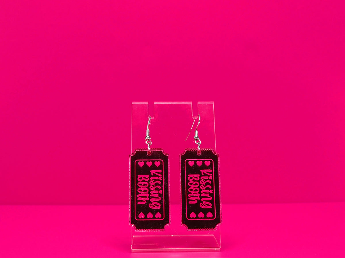 Mirrored pink kissing booth ticket earrings on a clear stand against a pink background.