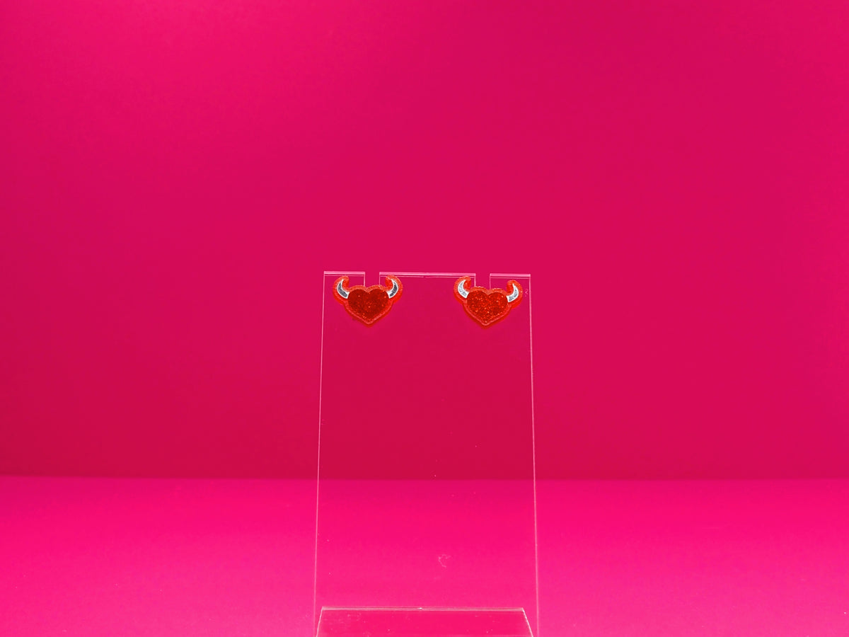 Devil horned heart earrings on a clear disay against a pink background.