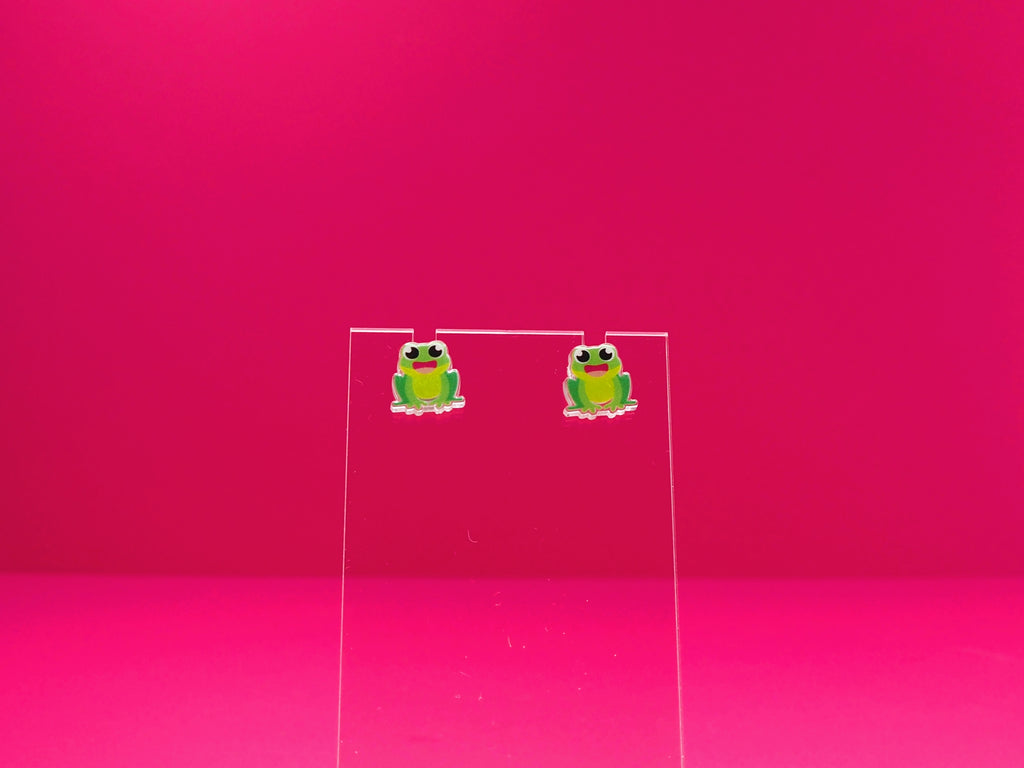 Frog earrings on a clear display against a pink background.