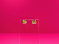 Frog earrings on a clear display against a pink background.