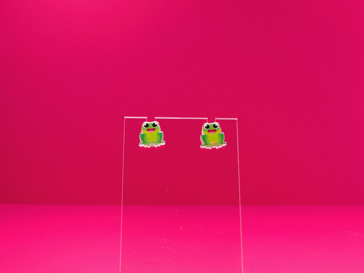 Frog earrings on a clear display against a pink background.