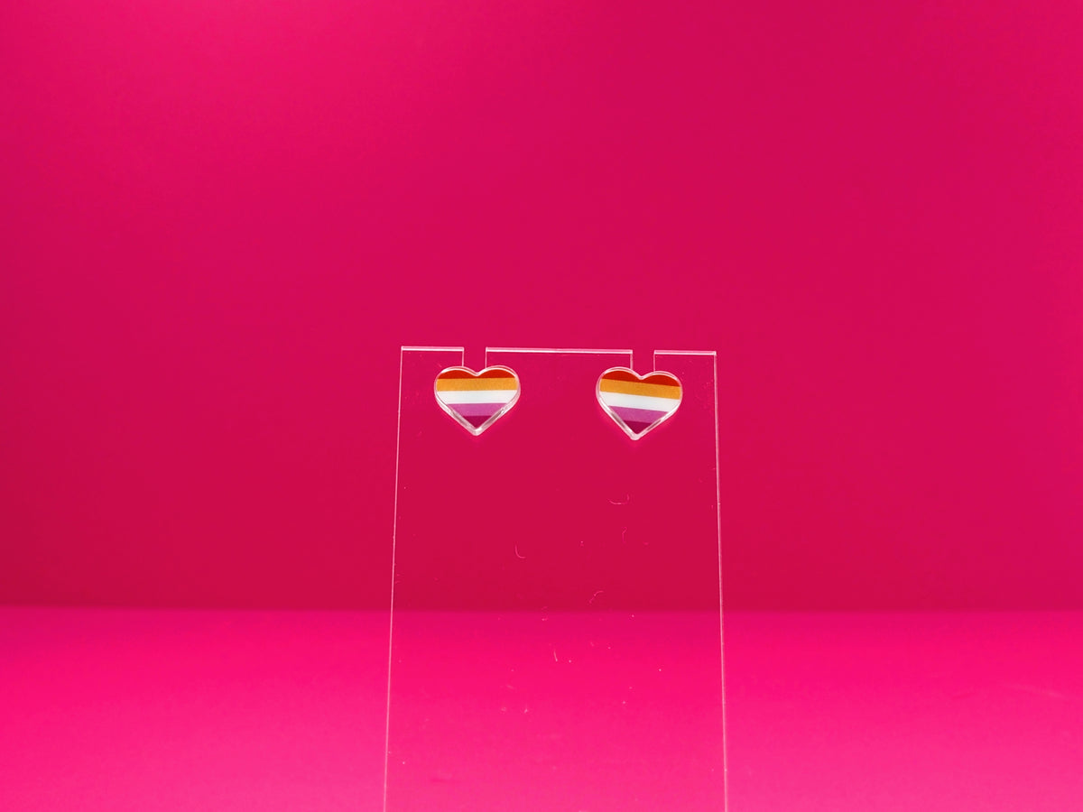 Lesbian pride heart earrings on a clear display against a pink background.