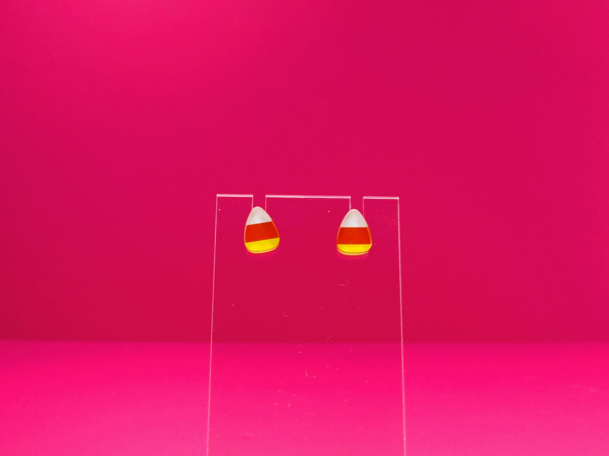 Candy corn earrings on a clear display against a pink background.