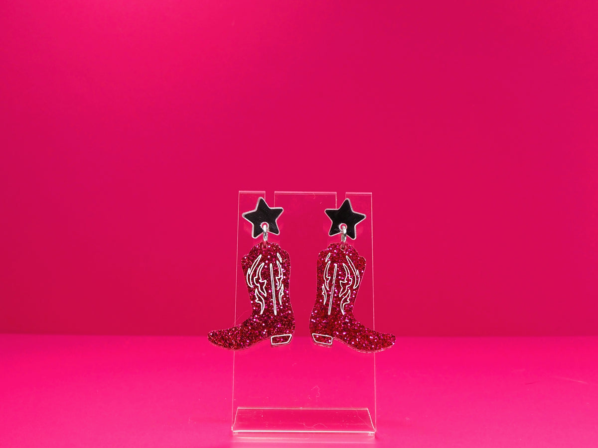 Pink cowboy boot earrings on a clear display against a pink background.