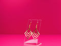 Gold geometric earrings on a clear display against a pink background.