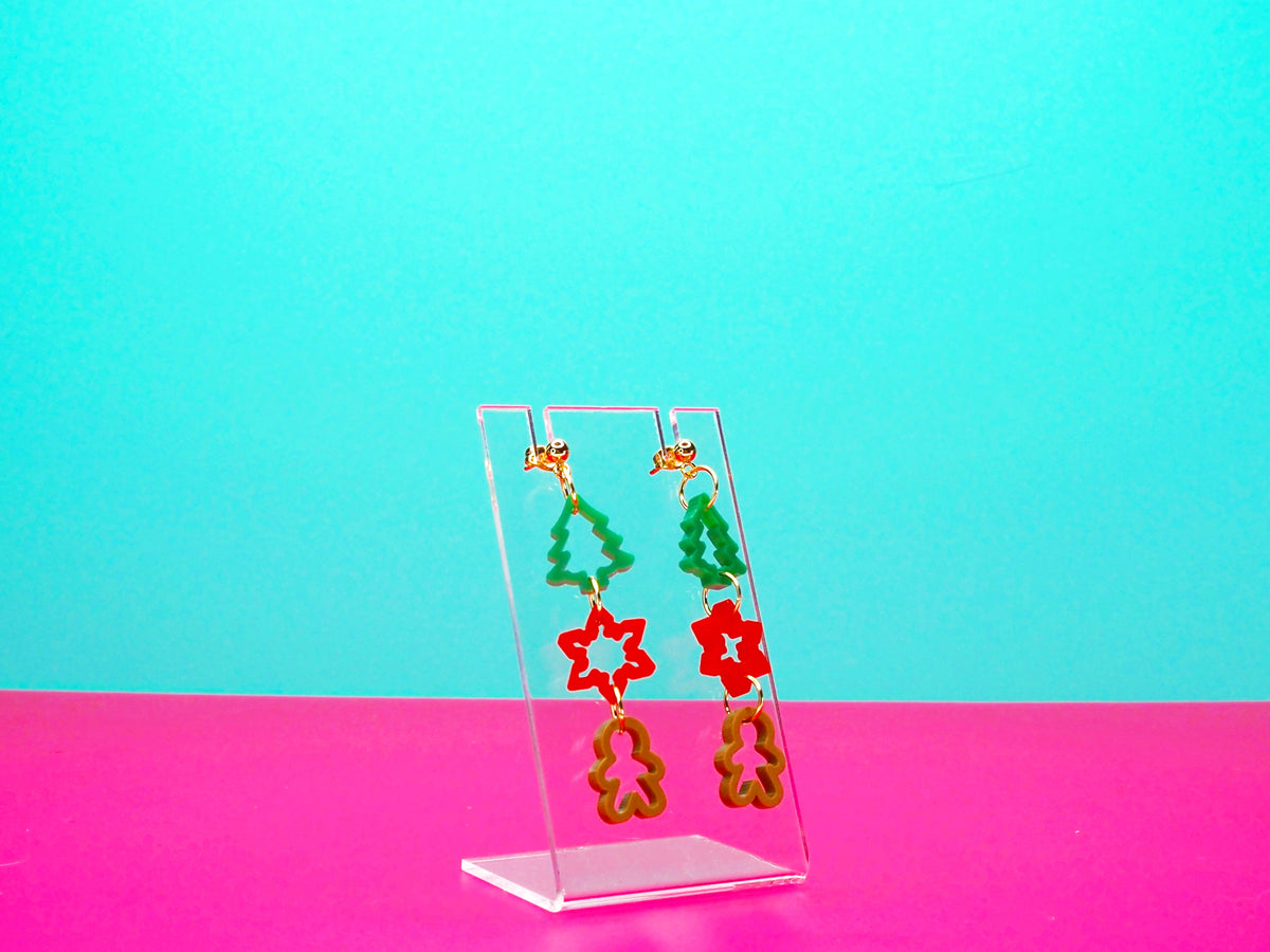 Christmas cookie cutter earrings on a clear display against a pink background.