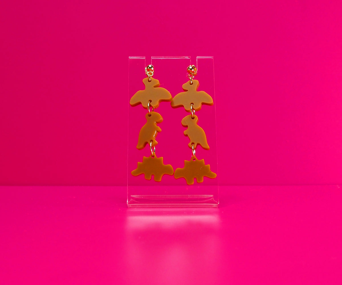 Dinosaur nugget earrings on a clear stand against a pink background.