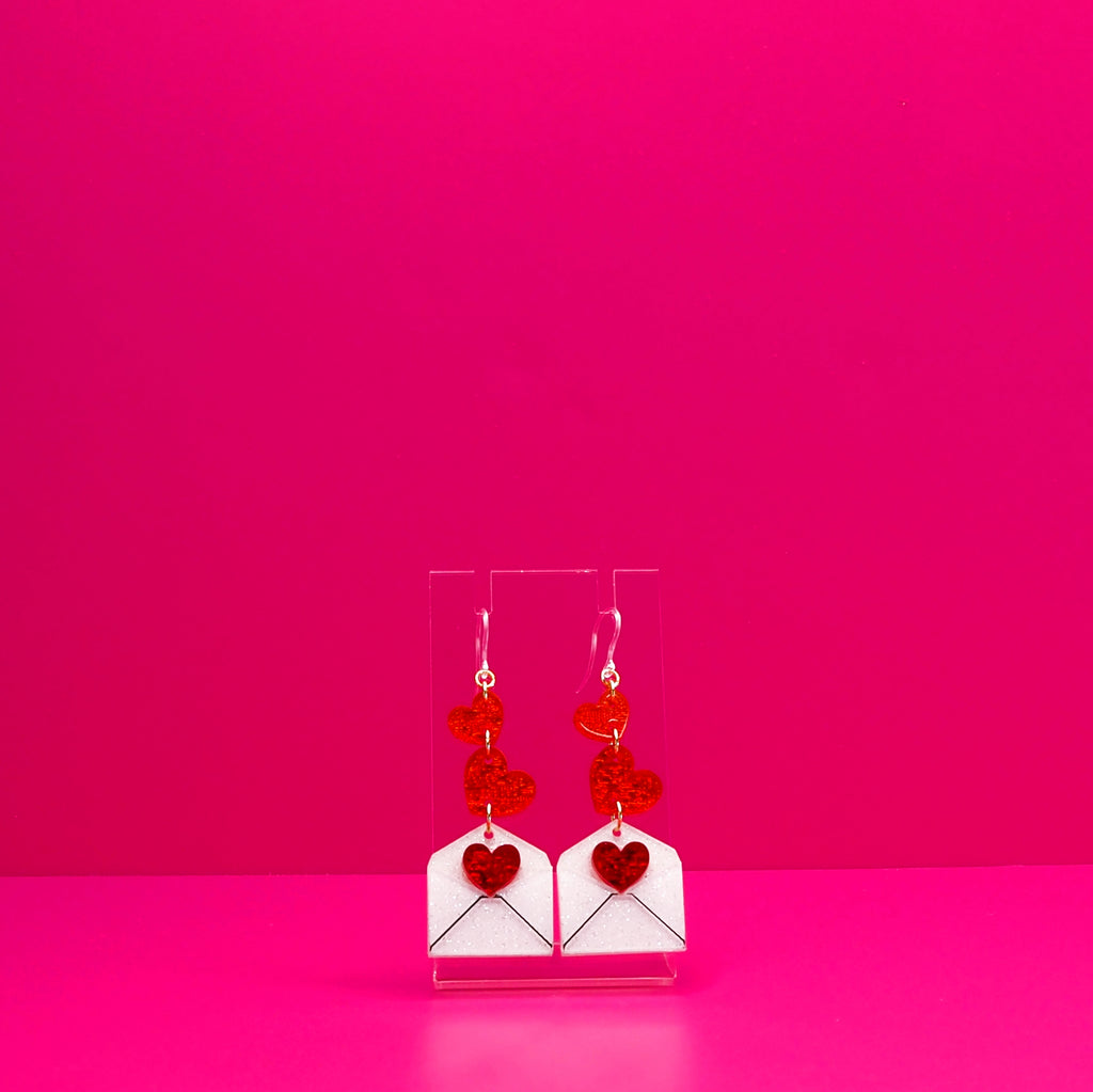 Red hearts cascading into a white envelope on a clear stand against a pink background.