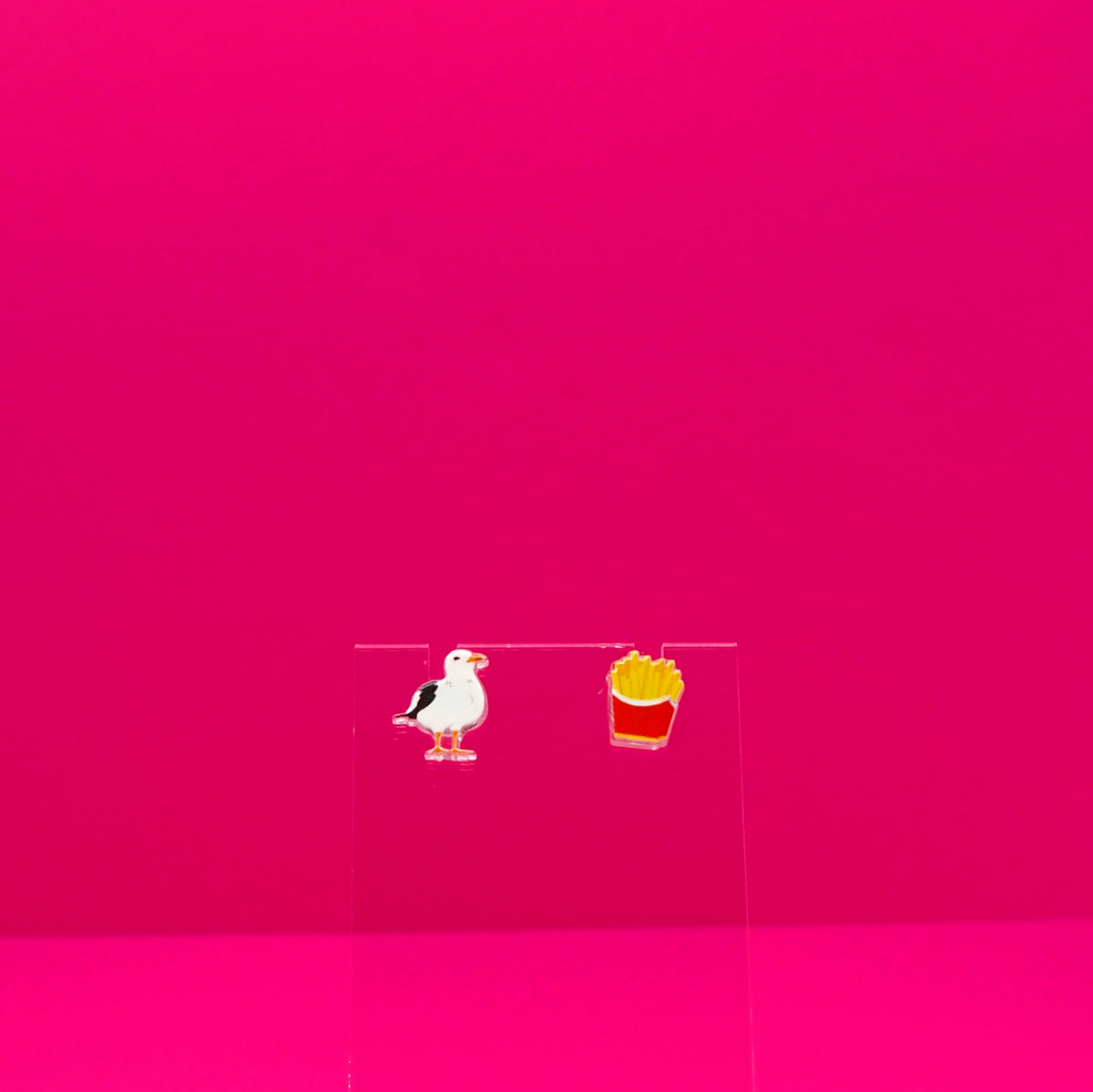 A seagull and a pack of French fries earrings on a clear stand against a pink background.