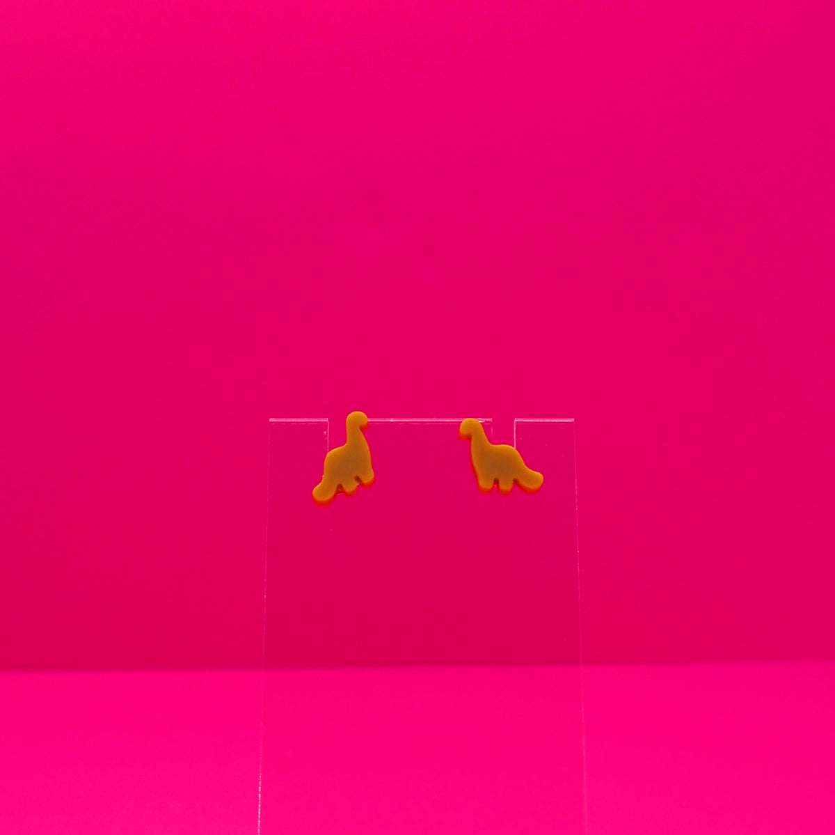 Brachiosaurus earrings on a clear stand against a pink background.