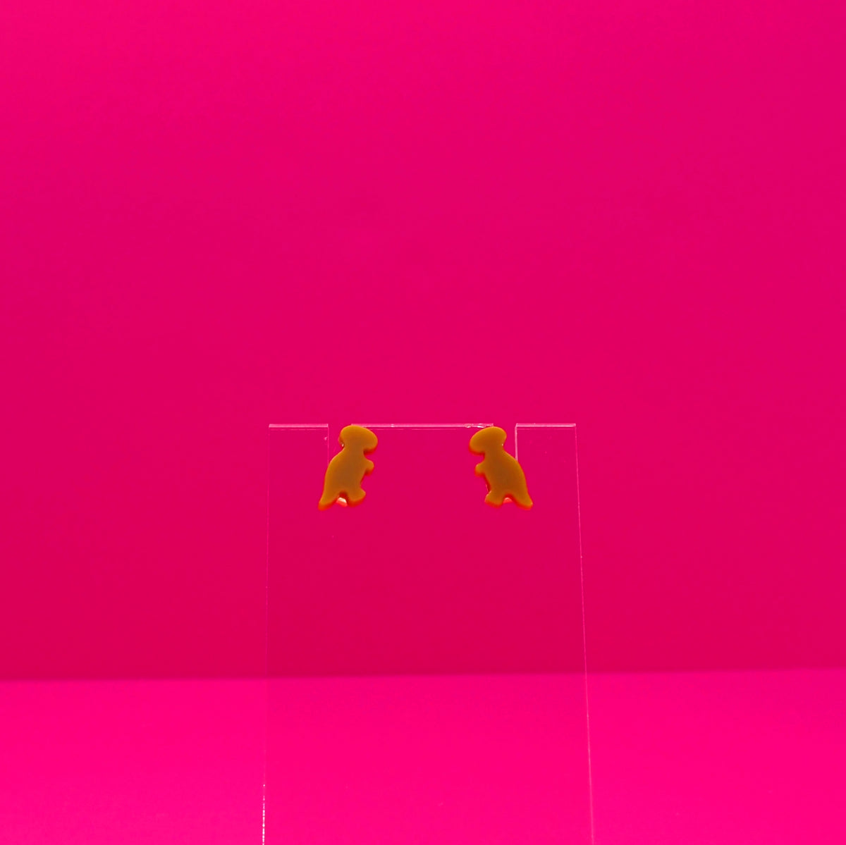 T-Rex earrings on a clear stand against a pink background.