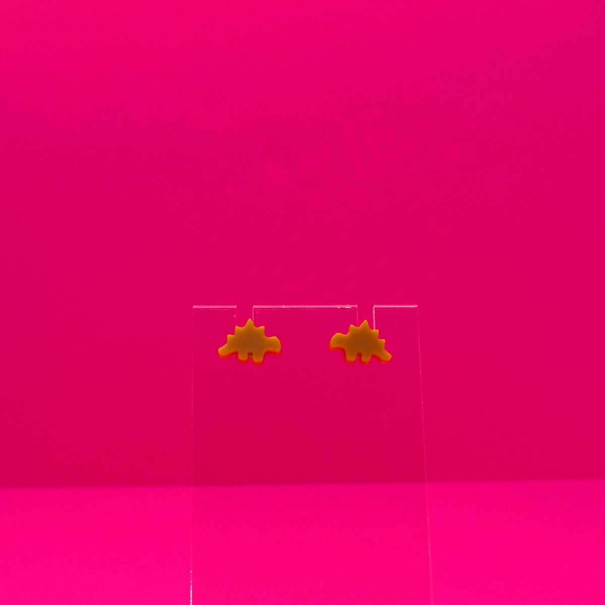 Stegosaurus earrings on a clear stand against a pink background.