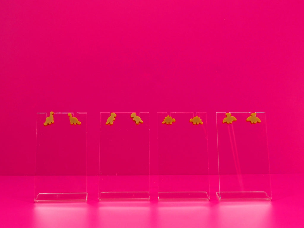 Set of brown dinosaur earrings on clear stands against a pink background