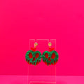 Red apple with green pearl goo earrings on a clear display against a pink background.