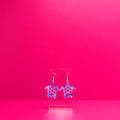 Purple  blue, pink, white floral sea turtle earrings on a clear display against a pink background.