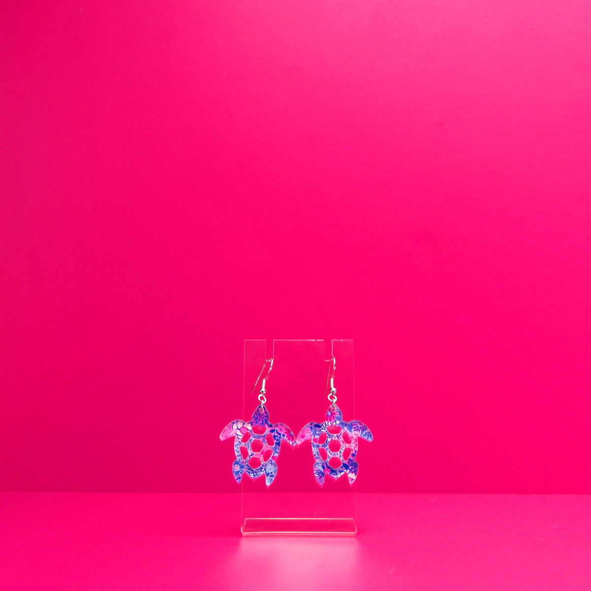 Purple  blue, pink, white floral sea turtle earrings on a clear display against a pink background.