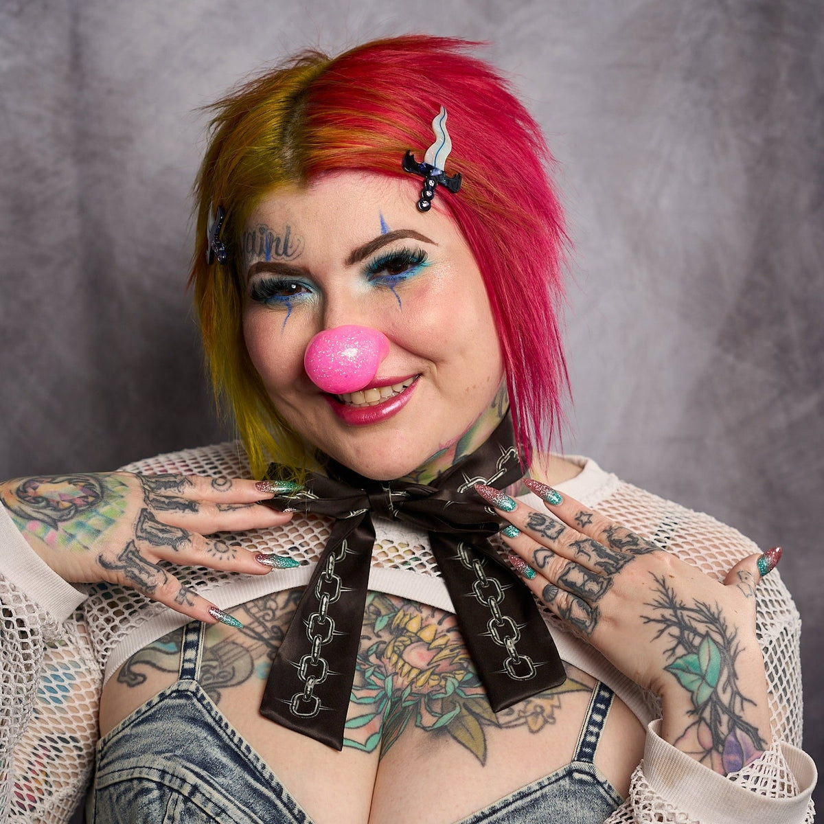 Model with split red and yellow hair wearing hair clip while wearing a pink clown nose against a dark background.