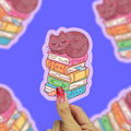 Brown cat sleeping on top of a stack of books. Books are pink, orange, yellow, green, and blue.