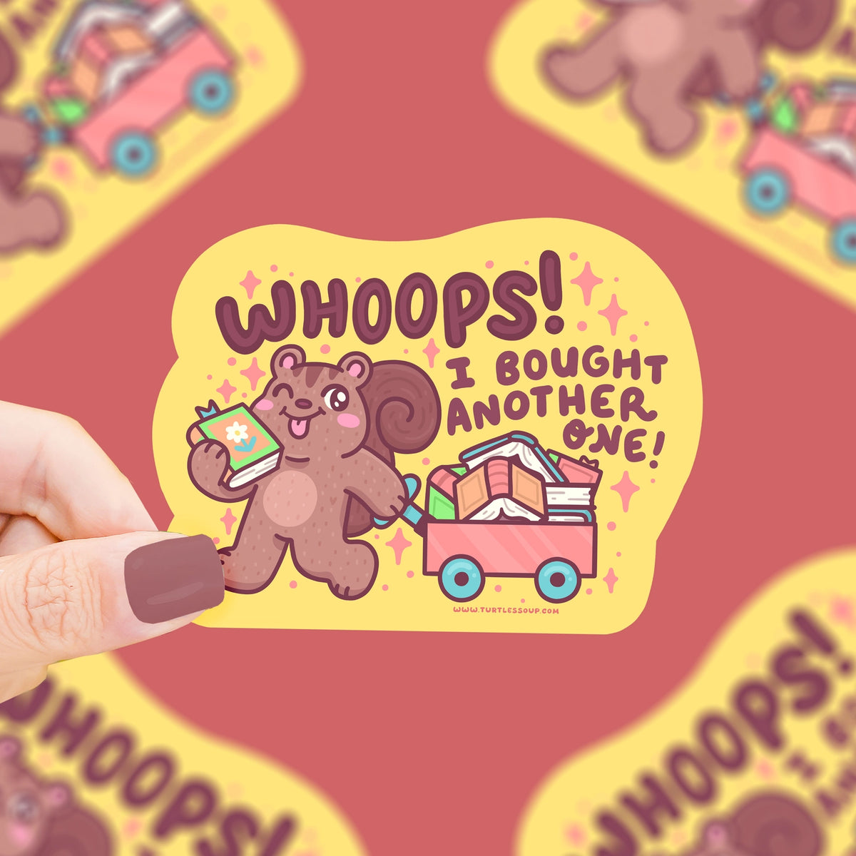 Hand holding a sticker with a brown squirrel pulling a red wagon filled with multi colored books in a pile against a yellow background surrounded by red diamond shapes. Sticker is against a red background with other copies of the sticker out of focus.