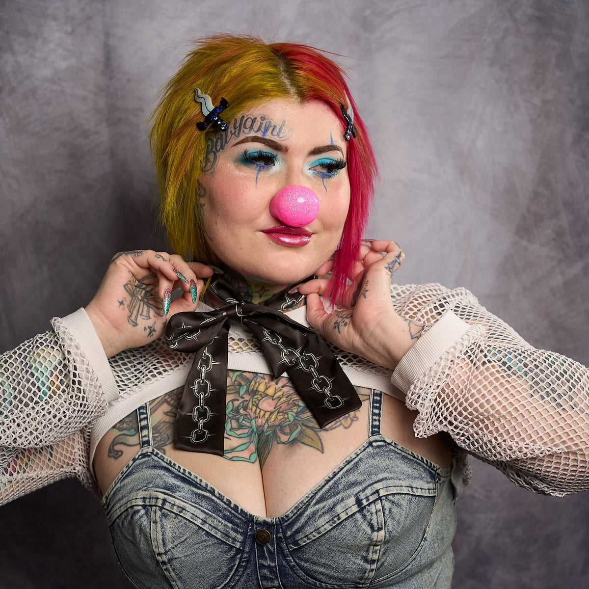 Model with split red and yellow hair wearing hair clip while wearing a pink clown nose against a dark background.