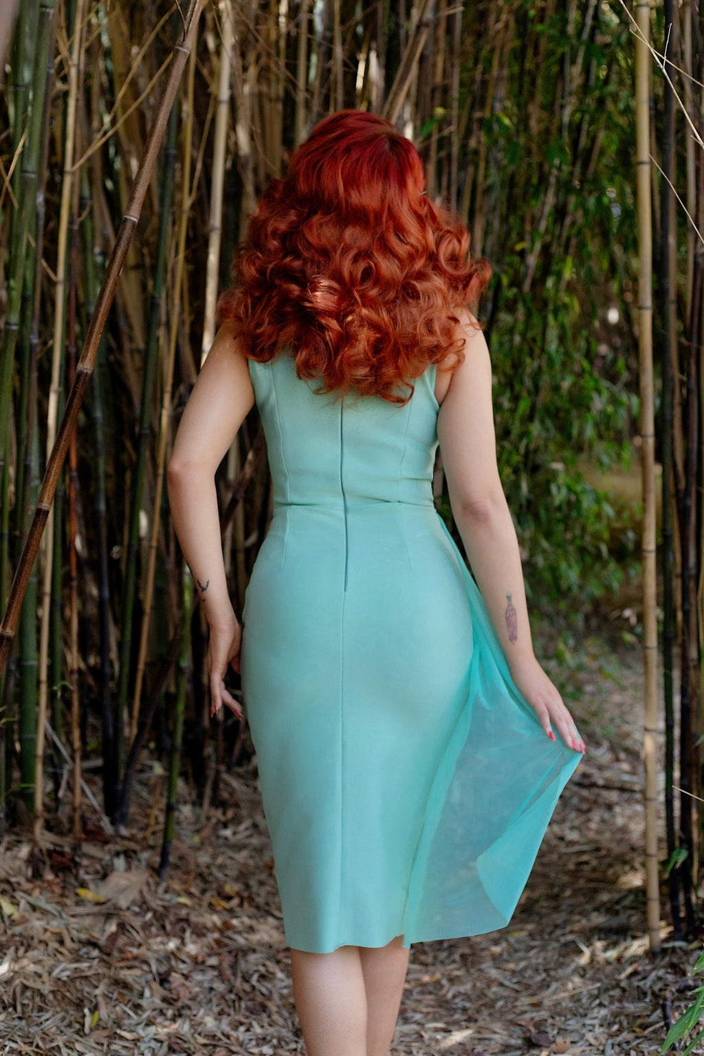 Model with bright red hair facing away to show back of the dress against a bamboo forest background