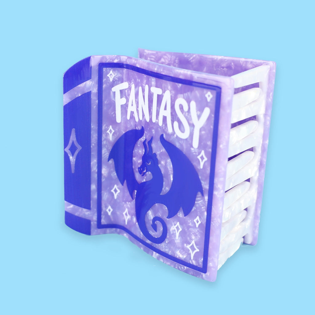 Purple book hair claw with a dark purple dragon on the front of the book with the word 'Fantasy' in white font. Hair claw is against a blue background.