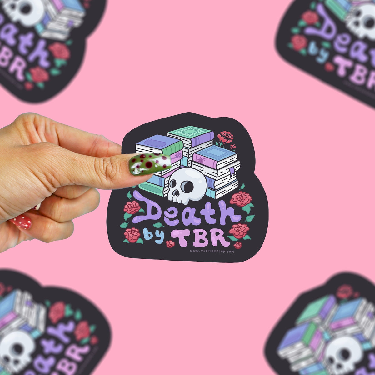 A hand with patterned nail polish holds a black die-cut sticker featuring a skull and books with the text 'Death by TBR' in purple and pink, against a pink background.