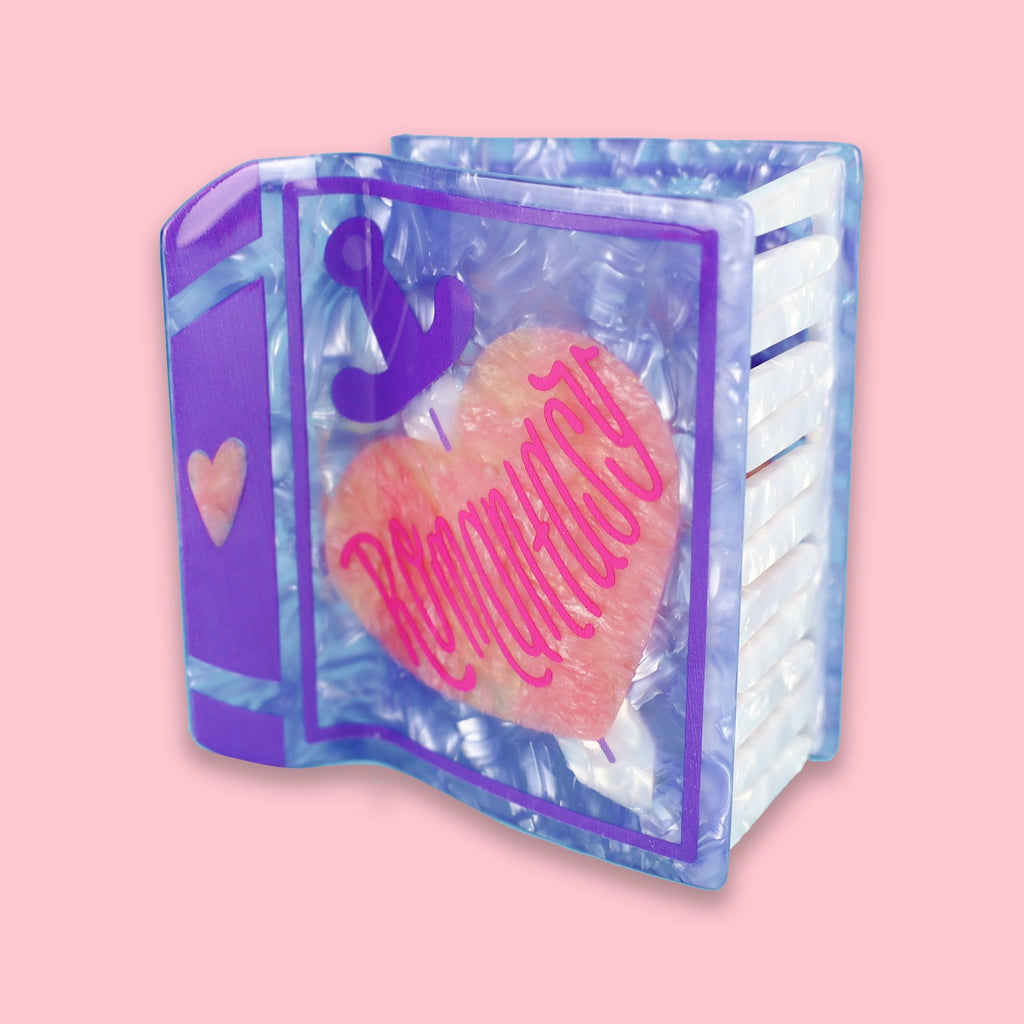 Marbled purple book hair claw with a silver sword going through a peach colored heart and the phrase 'Romantasy' in red lettering. Hair claw is against a pink background.