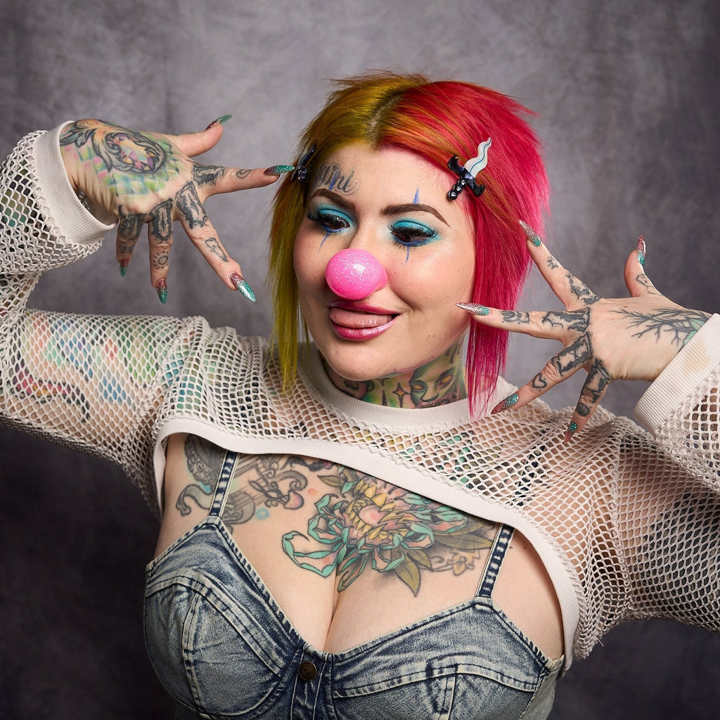 Model with split red and yellow hair wearing hair clip while wearing a pink clown nose against a dark background.