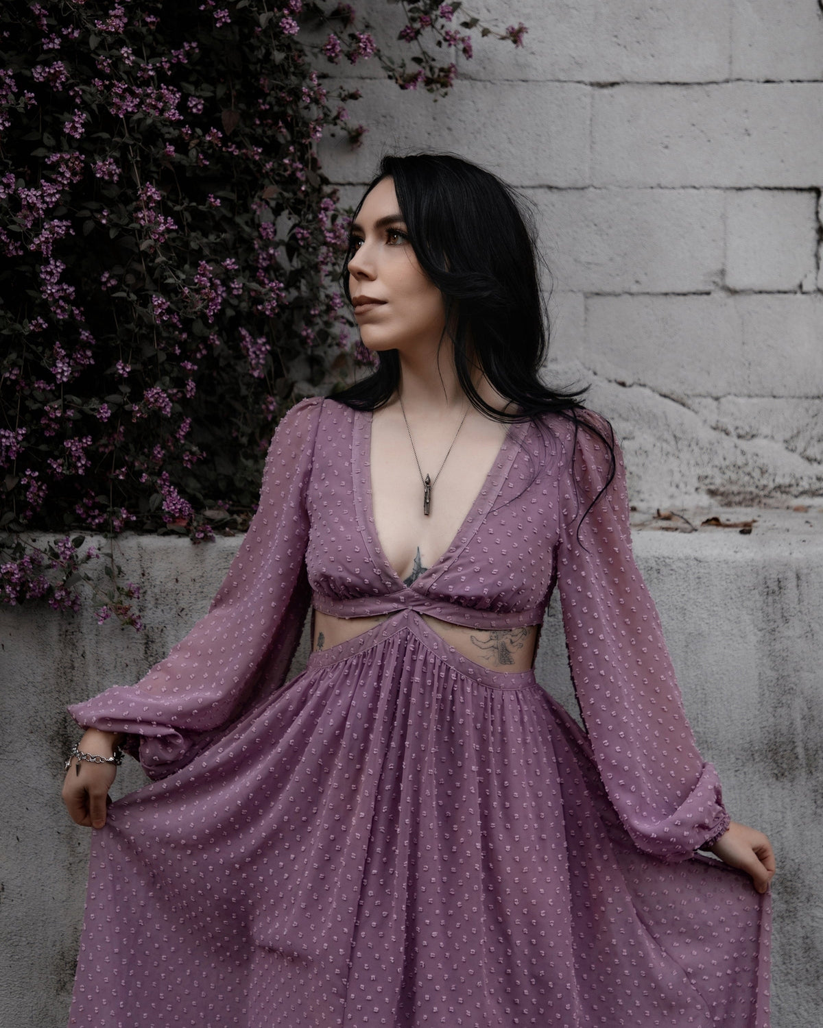 Female presenting model in the purple Lunara dress standing against a stone wall with purple flowers.