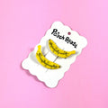 Two ripe banana clips on a white hang card against a pink background.