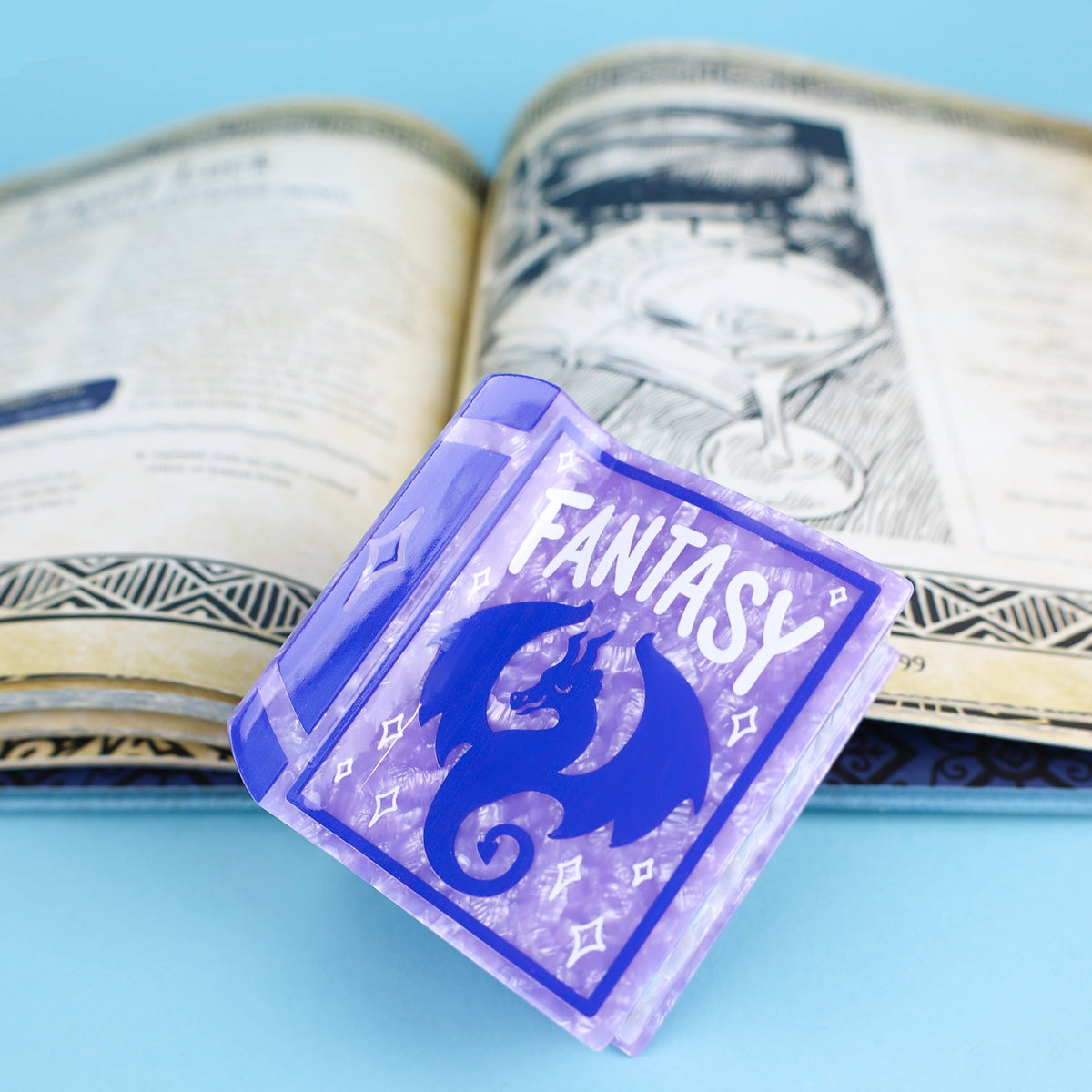 Purple book hair claw with a dark purple dragon on the front of the book with the word 'Fantasy' in white font. Hair claw is resting on an open book against a blue background.