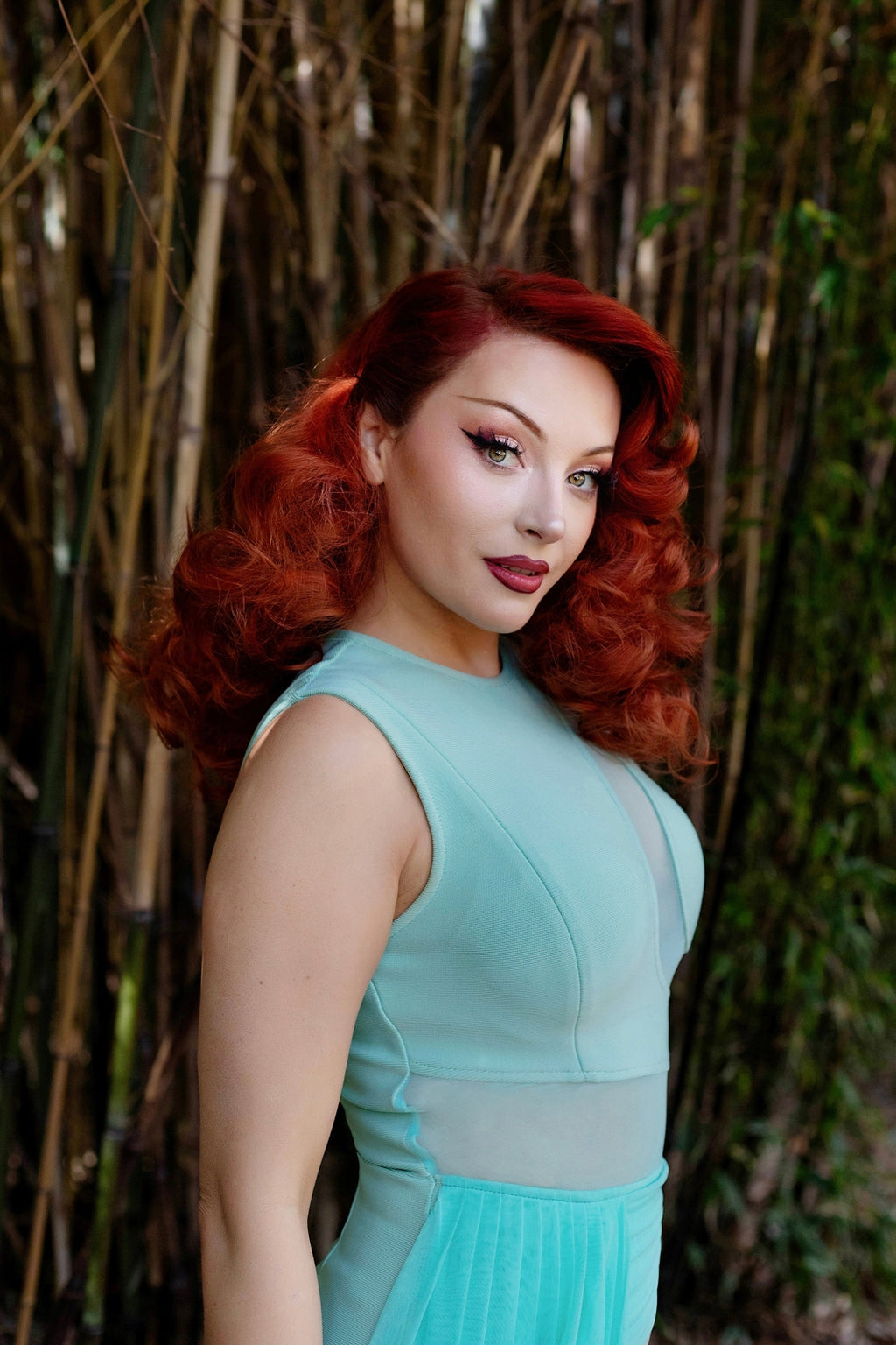 Portrait of model with bright red hair facing to the right against a bamboo background.